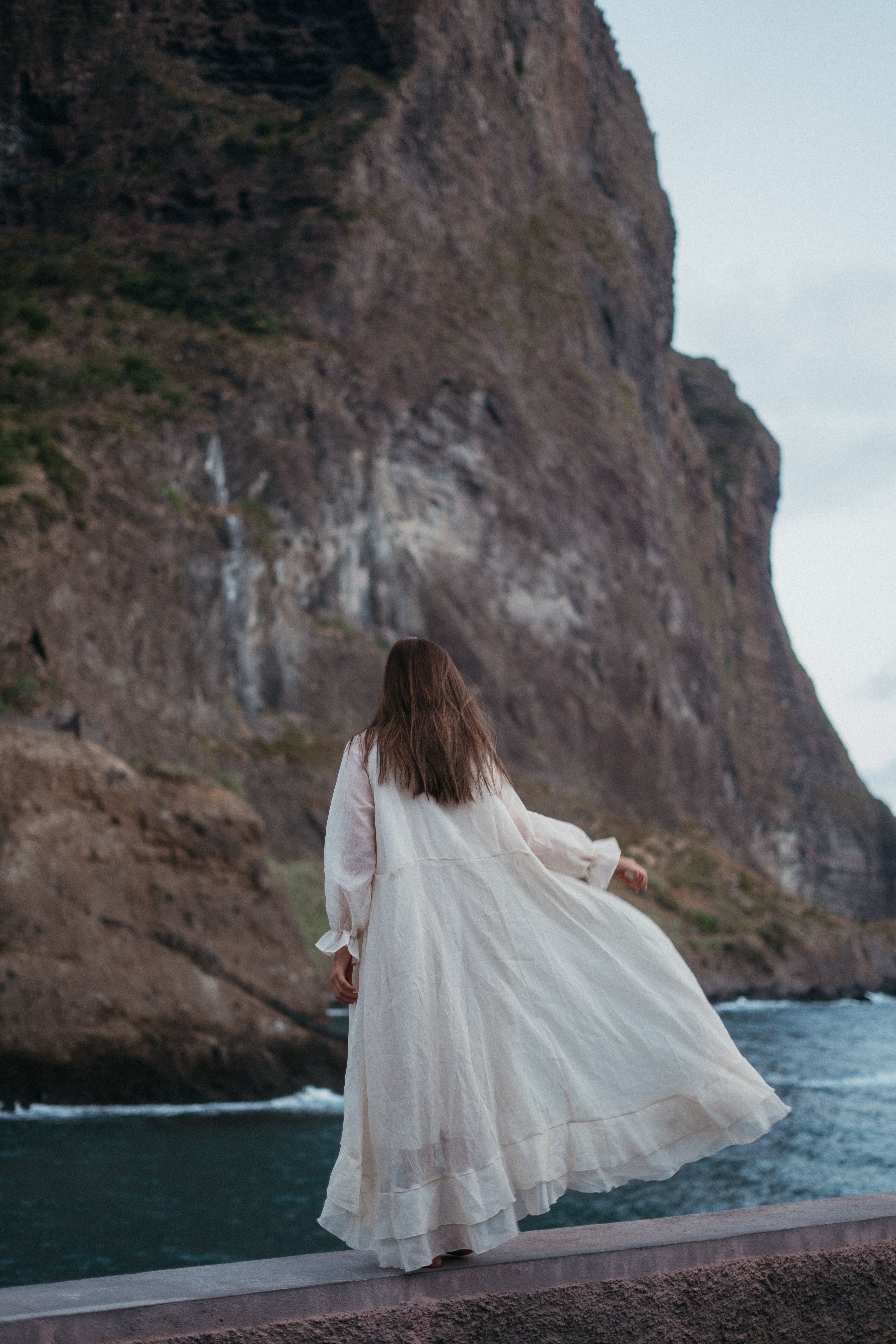 Woman in a white dress standing by the sea beneath a tall cliff
