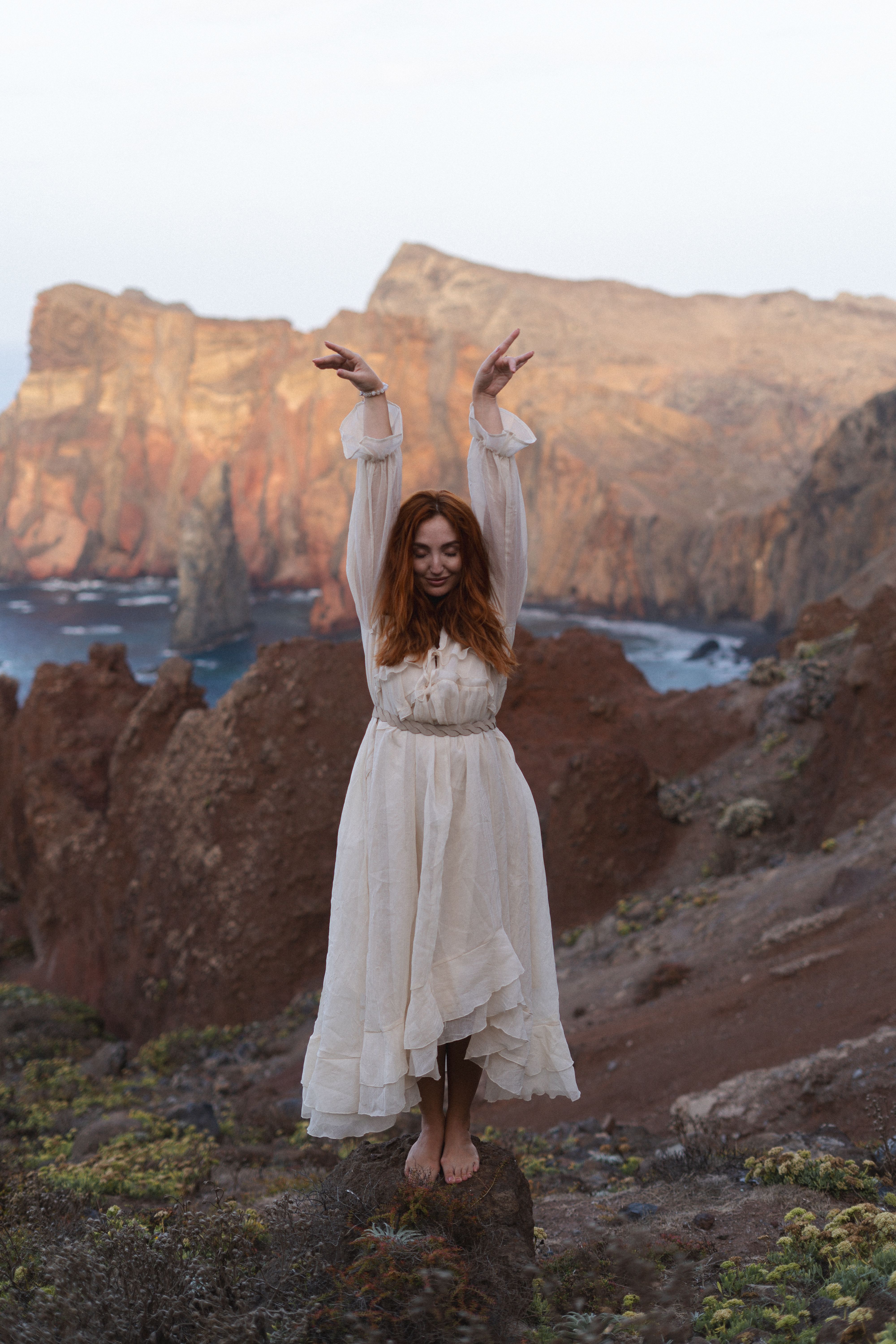 Woman in a white dress raising her arms against a rocky coast