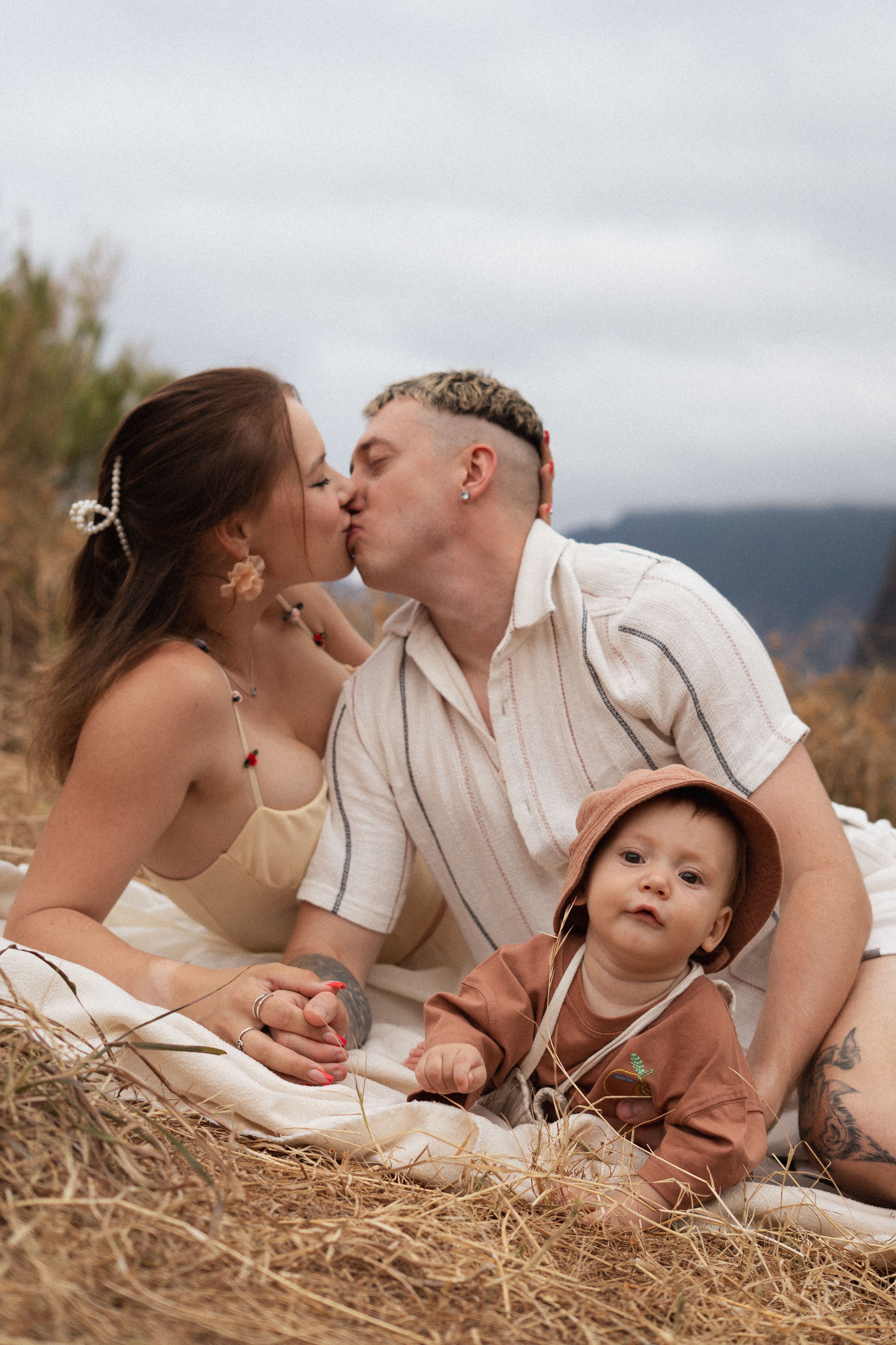 Parents kissing beside their baby on a blanket in tall grass